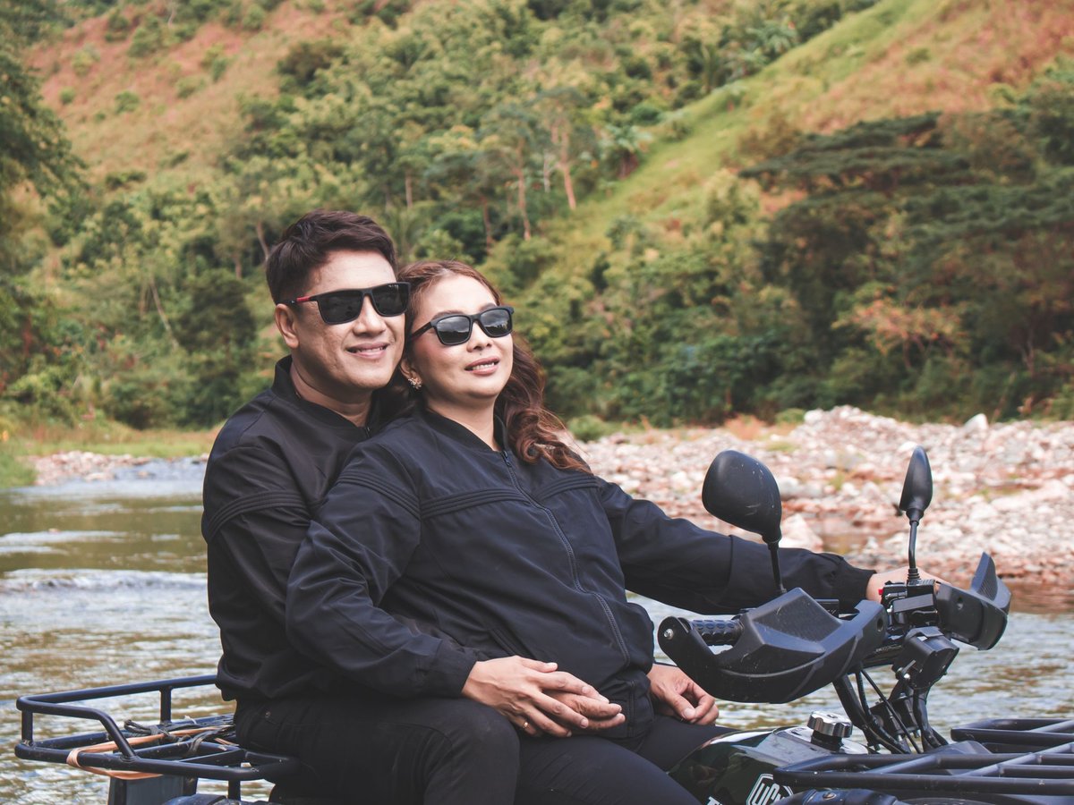 Couple riding an ATV through a shallow rocky stream with a green hillside behind them